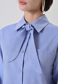 Camicia azzurro chiaro con un fiocco annodato al collo, chiusura con bottoni e taschino sul petto. Il tessuto ha una leggera trama.