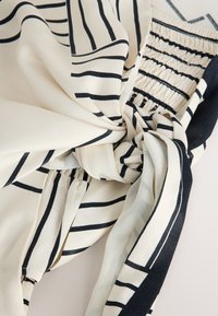 Tissu beige avec des rayures géométriques noires, détail noué et section élastique froncée, montrant une texture douce et un motif en gros plan.