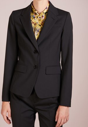 Blazer noir ajusté à deux boutons porté sur une chemise jaune à col à motif floral et un pantalon noir assorti.