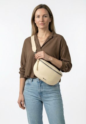 Vrouw met een bruin shirt en lichtblauwe spijkerbroek houdt een crèmekleurige, getextureerde crossbodytas met een ritssluiting en brede band over haar borst vast.