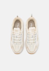 Light beige athletic sneakers with mesh panels, textured accents, and white laces; features a small pull tab and Fila branding on the tongue.