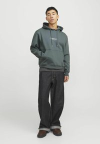Dark green hoodie with a kangaroo pocket, drawstrings, and logo; paired with dark blue wide-leg jeans and brown shoes.