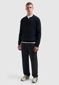 Jonge man in een zwarte polo met lange mouwen, zwarte broek en zilveren sneakers tegen een effen lichte achtergrond.