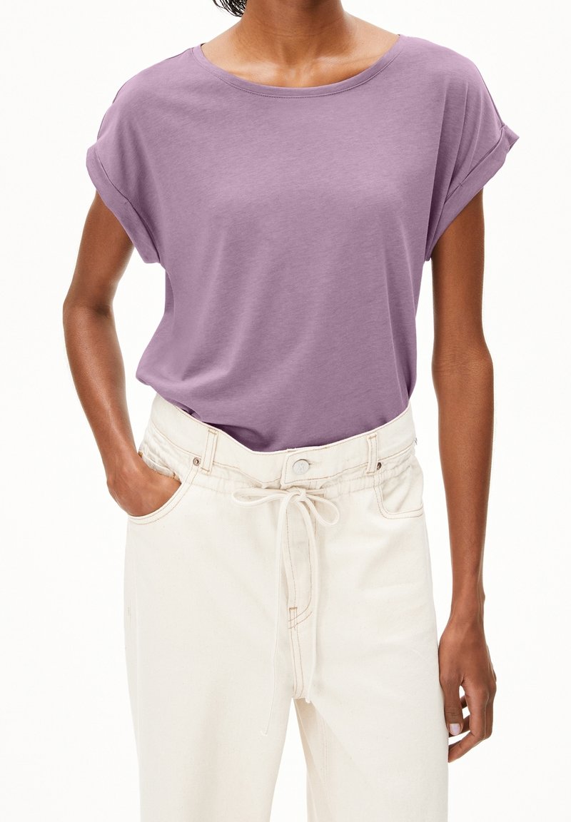 Femme portant un haut lilas à manches courtes rentré dans un pantalon taille haute écru avec une ceinture à cordon, main dans la poche.