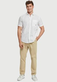 Witte linnen shirt met korte mouwen en een borstzak, gecombineerd met beige broek met trekkoord en witte sneakers, op een neutrale achtergrond.