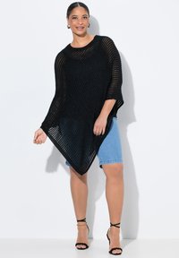 Svart stickad poncho med en lös design och öppen väv, med breda ärmar och en spetsig vågkant. Bärs över denimshorts och svarta sandaler.