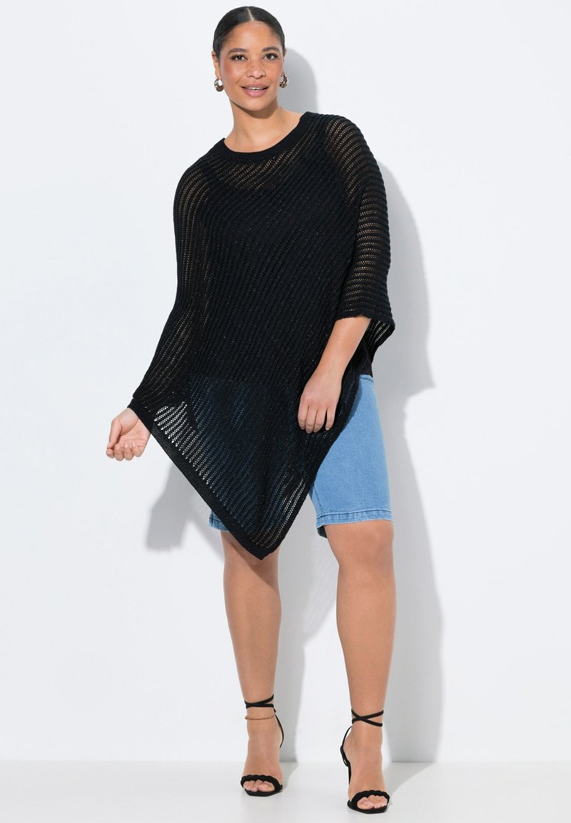 Svart stickad poncho med en lös design och öppen väv, med breda ärmar och en spetsig vågkant. Bärs över denimshorts och svarta sandaler.