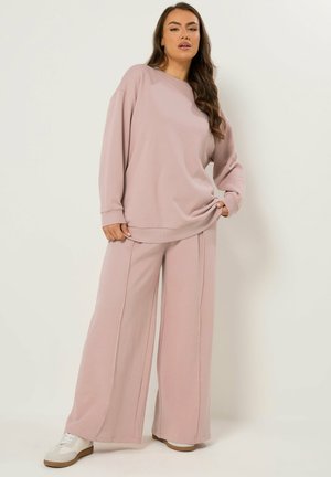 Vrouw staat, draagt een loszittende licht mauve lange mouwen top en wijde broek met witte sneakers, tegen een effen lichte achtergrond.