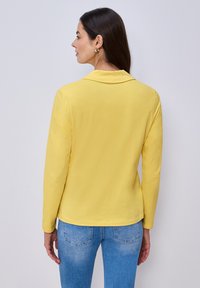 Femme aux longs cheveux bruns portant un haut jaune à manches longues et un jean bleu, debout de profil face à droite sur un fond clair uni.
