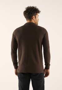 Moški, obrnjen stran, oblečen v temno rjavo srajco z dolgimi rokavi in črne kavbojke, s preprostim svetlim ozadjem.