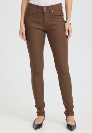 Pantalon slim marron avec deux boutons sur le devant et poches, porté avec des ballerines noires pointues et un haut blanc, visible de la taille jusqu'aux pieds.