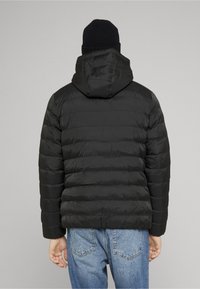 Doudoune noire avec coutures horizontales, capuche attachée et poignets élastiques, portée avec un jean bleu clair ; design minimaliste.