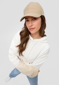 Casquette beige avec une visière courbée et un texte embossé discret. Le modèle porte un hoodie blanc avec des accents beiges et un jean bleu, sur un fond gris.