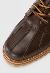 Brown leather shoe featuring a rounded toe, stitched detailing, two-tone laces, and a textured sole with a contrasting light brown rim.