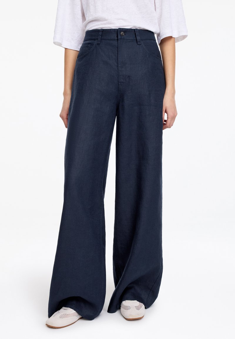 Femme portant un pantalon bleu foncé taille haute à jambes larges avec poches avant, associé à des baskets blanches et un top blanc ample à manches courtes.
