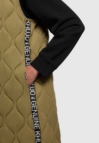 Gilet verde oliva trapuntato con tasche laterali, caratterizzato da una superficie testurizzata e accenti neri con branding bianco lungo la zip.