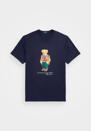 Maglietta blu navy con un orso che indossa un blazer marrone, un maglione viola, pantaloni verdi e scarpe marroni, con la scritta "Polo Bear by Ralph Lauren" sotto.