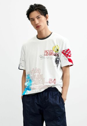 Joven con cabello oscuro que lleva puesta una camiseta blanca con estampados en rojo, azul y amarillo y pantalones negros, de pie con las manos en los bolsillos.