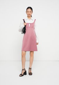 Een roze jurk met een getailleerde bodice, vierkante halslijn en sleutelgatdetail, gecombineerd met een witte blouse en een zwarte handtas, gedragen met zwarte sandalen met bandjes.
