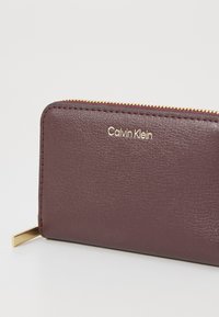 Burgundy leather wallet with zip closure, gold hardware, and embossed logo. Features a textured surface and a sleek rectangular shape.