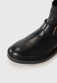Botín negro de cuero hasta el tobillo con puntera redondeada, costuras decorativas y una correa con hebilla lateral sobre un fondo blanco.