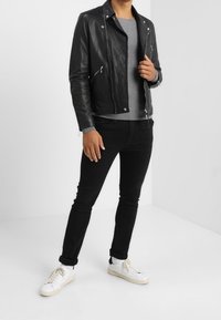 Black leather jacket with silver hardware, grey sweater underneath, black jeans, and white sneakers. Modern fit with zippers on the jacket.