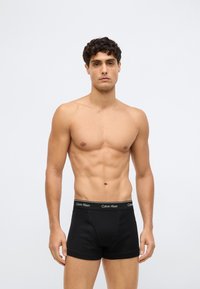 Homem sem camisa com cabelo curto e cacheado, vestindo boxers pretos da Calvin Klein, em pé contra um fundo branco liso.