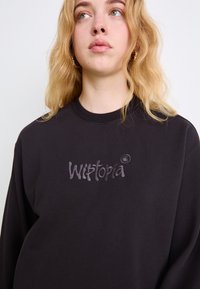 Czarna bluza z bawełny, o luźnym kroju i okrągłym dekolcie. Zauważalne logo "Wistoria" w kontrastującym odcieniu szarości na piersi.