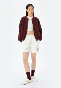 Bordowa kurtka bomberka na zamek, noszona na białej bluzce crop top, zestawiona z białymi szortami z logo i bordowymi skarpetkami, uzupełniona o białe sneakersy.