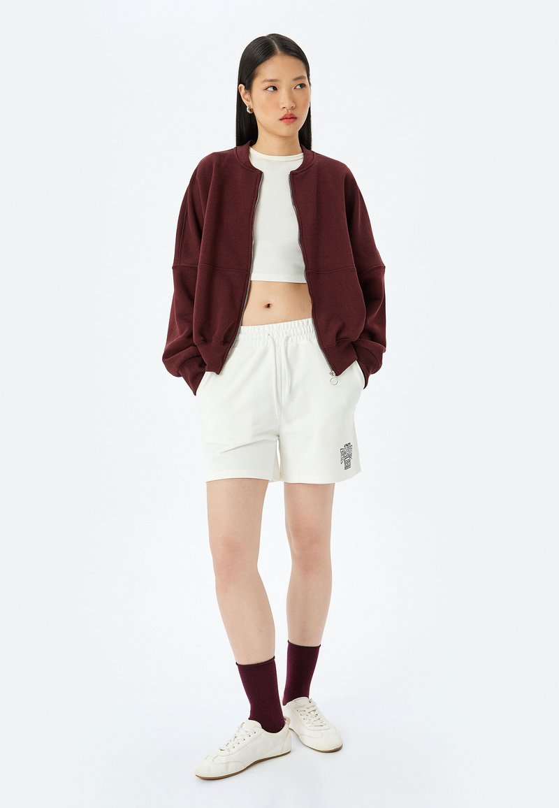 Bordowa kurtka bomberka na zamek, noszona na białej bluzce crop top, zestawiona z białymi szortami z logo i bordowymi skarpetkami, uzupełniona o białe sneakersy.