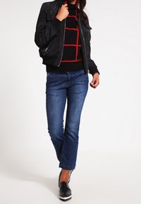 Person in einer schwarzen Bomberjacke, einem schwarz-roten karierten Pullover, blauen geraden Jeans und schwarzen Plateauschuhen.