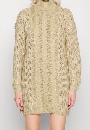 Beige chunky gebreide trui-jurk met kabelpatroon en col, met lange mouwen, gedragen door een persoon met zichtbare handen en benen.