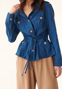 Blå linnekavaj med knäppta fickor, midjan framtvingad med snodd, och dragsko. Har krage och träknappar. Lättviktig textur.