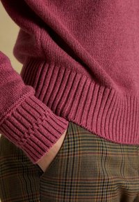 Roosa kootud svetter soonikute ja alläärega, kombineeritud ruuduliste pükstega, millel on pruunide, kollaste ja roheliste toonide segu.