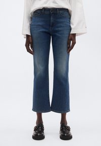 Sötétkék farmer rövidített, bővülő szabással. Világos mosott hatású és apró szegecsekkel díszített szegélyekkel. Fekete cipővel viselve.