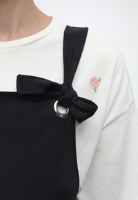 Λευκό πουκάμισο με μικρή ροζ κεντητή τριανταφυλλιά, φορεμένο κάτω από μαύρη σαλοπέτα με ιμάντα ώμου δεμένο σε φιόγκο, που έχει ασημένιο μεταλλικό κρίκο.