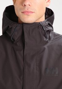 Waterproof dark brown hooded jacket with adjustable drawstrings, mesh lining, and a subtle logo accent on the front.