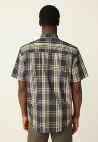 Košeľa s krátkym rukávom v károvanej vzorke v námorníckej modrej, krémovej a hnedej farbe. Má zapínací golier a zadný diel s yoke, textúrovanú tkaninu a rovný lem.