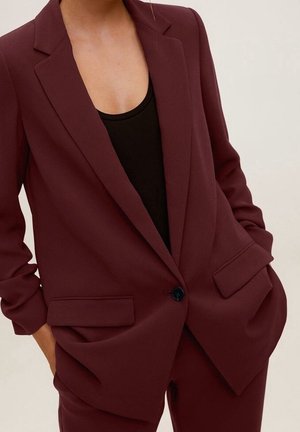 Femme portant un blazer ajusté bordeaux foncé sur un haut noir, les mains dans les poches, visible de la moitié de la poitrine à la moitié de la cuisse.