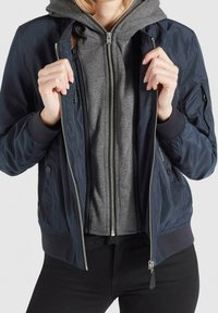 Marinblå bomberjacka med ribbade muddar, dragkedja fram och sidofickor, lagd över en grå zip-up hoodie. Slät, lätt material.