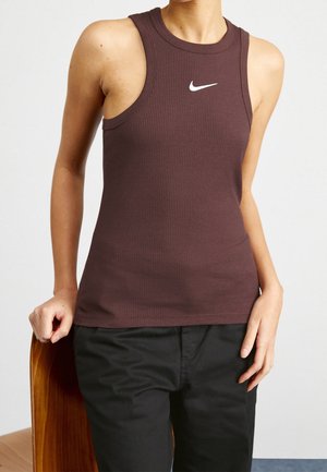 Personne portant un débardeur côtelé sans manches marron foncé avec un logo Nike blanc, pantalon noir, debout avec une main posée sur une chaise en bois.