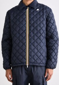 Giacca trapuntata blu navy con colletto; presenta dettagli della zip a strisce gialle e bianche e un piccolo logo sul petto sinistro. Texture morbida e imbottita.
