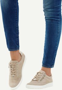 Béžové semišové tenisky s bílými gumovými podrážkami, zdobené zipem a šňůrkami, v kombinaci s krátkými modrými džínami.