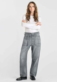 Pantalons en denim gris à jambe large avec plusieurs poches et des coutures renforcées, associés à un haut ajusté à manches longues blanc et des chaussures noires.