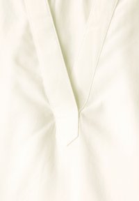 Blouse en ivoire avec un décolleté en V, tissu doux et détails angulaires sur le col. Texture lisse avec des accents de coutures subtiles.