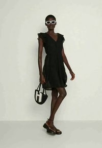 Μαύρο φόρεμα με φουσκωτά μανίκια και V λαιμόκοψη, συνδυασμένο με μια υφασμάτινη τσάντα σε μαύρο και λευκό. Το μοντέλο φοράει λευκά γυαλιά ηλίου και μαύρες σαγιονάρες.