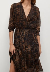 Mujer vistiendo un vestido envolvente de patrón marrón y negro con mangas tres cuartos y escote en V, sosteniendo la falda de un lado.