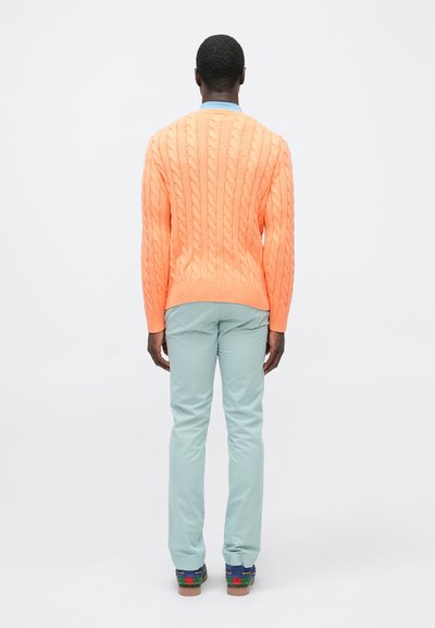 Φωτεινό πορτοκαλί πουλόβερ πλεκτό με στρογγυλή λαιμόκοψη, συνδυασμένο με ανοιχτό πράσινο παντελόνι και πολύχρωμα παπούτσια. Απλός, υφασμάτινος σχεδιασμός.