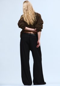 Oversized brun sweater og løse sorte bukser med en glat tekstur og vide ben, vist bagfra mod en lys baggrund.