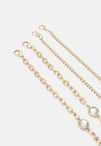 Quatre rallonges de bracelet en chaîne dorée avec fermoirs mousquetons, dont deux ornées de pierres précieuses circulaires transparentes, disposées côte à côte sur fond blanc.